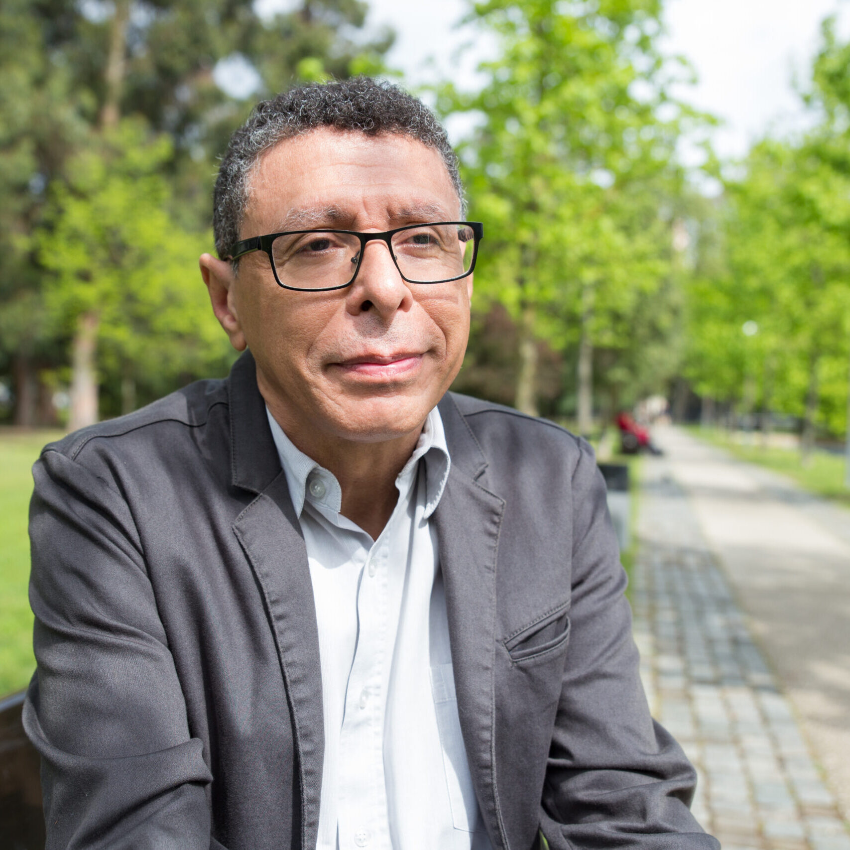 Relaxed middle-aged man sitting on bench in city park. Guy wearing casual clothes and relaxing with walkway and green trees in background. Relaxation and nature concept. Front view.